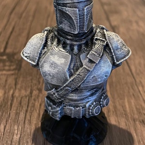 May include: A 3D printed bust of a Mandalorian character from the Star Wars universe. The bust is painted in a silver color and features intricate details of the character's armor and helmet.