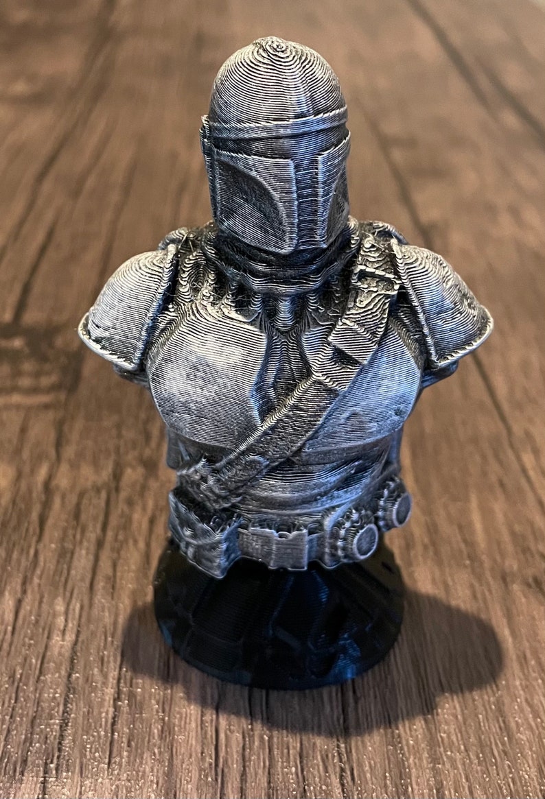 May include: A 3D printed bust of a Mandalorian character from the Star Wars universe. The bust is painted in a silver color and features intricate details of the character's armor and helmet.