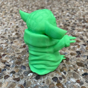 May include: A 3D-printed, light green figurine of a character with large ears and a robe. The figurine is posed with its arms outstretched. The figure is standing on a surface of small, multicolored stones.