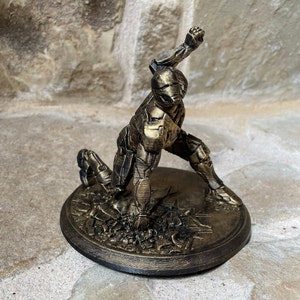 May include: A gold-painted figurine of a robot in a dynamic pose, standing on a circular base with a textured surface. The robot has a detailed design and appears to be made of plastic.
