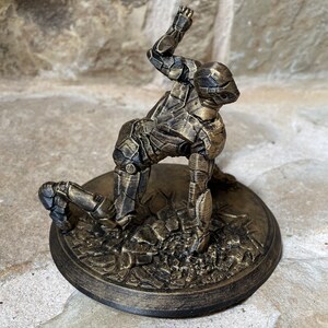 May include: A gold-painted figurine of a robot in a kneeling pose, with one arm raised above its head. The figurine is on a round, gold-painted base with a textured surface.
