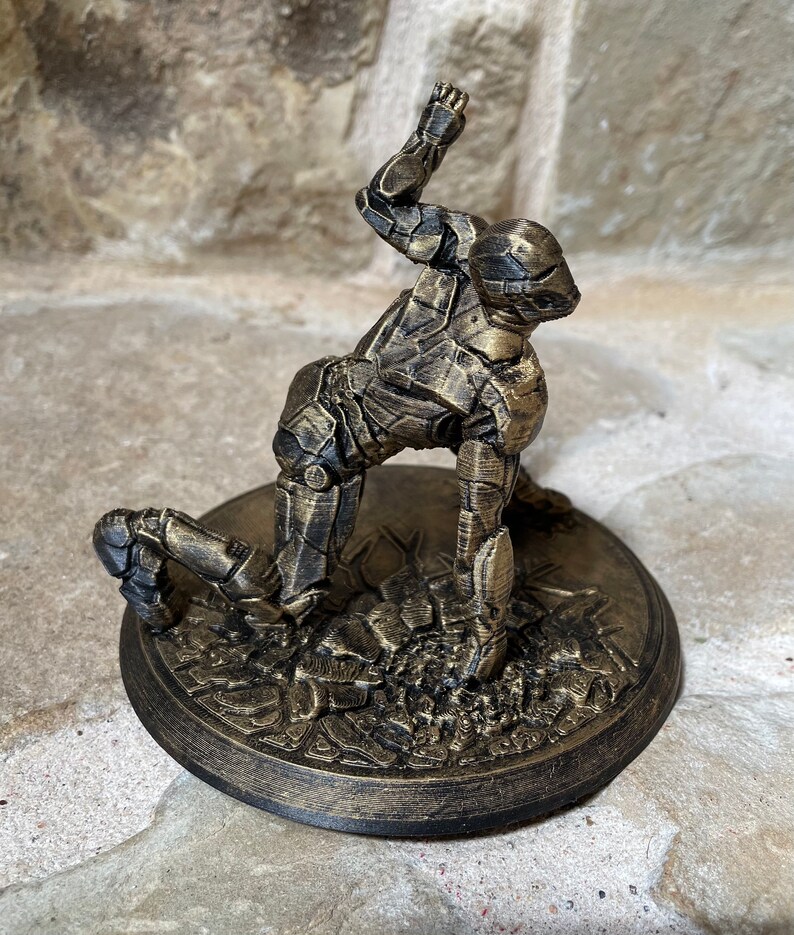 May include: A gold-painted figurine of a robot in a kneeling pose, with one arm raised above its head. The figurine is on a round, gold-painted base with a textured surface.