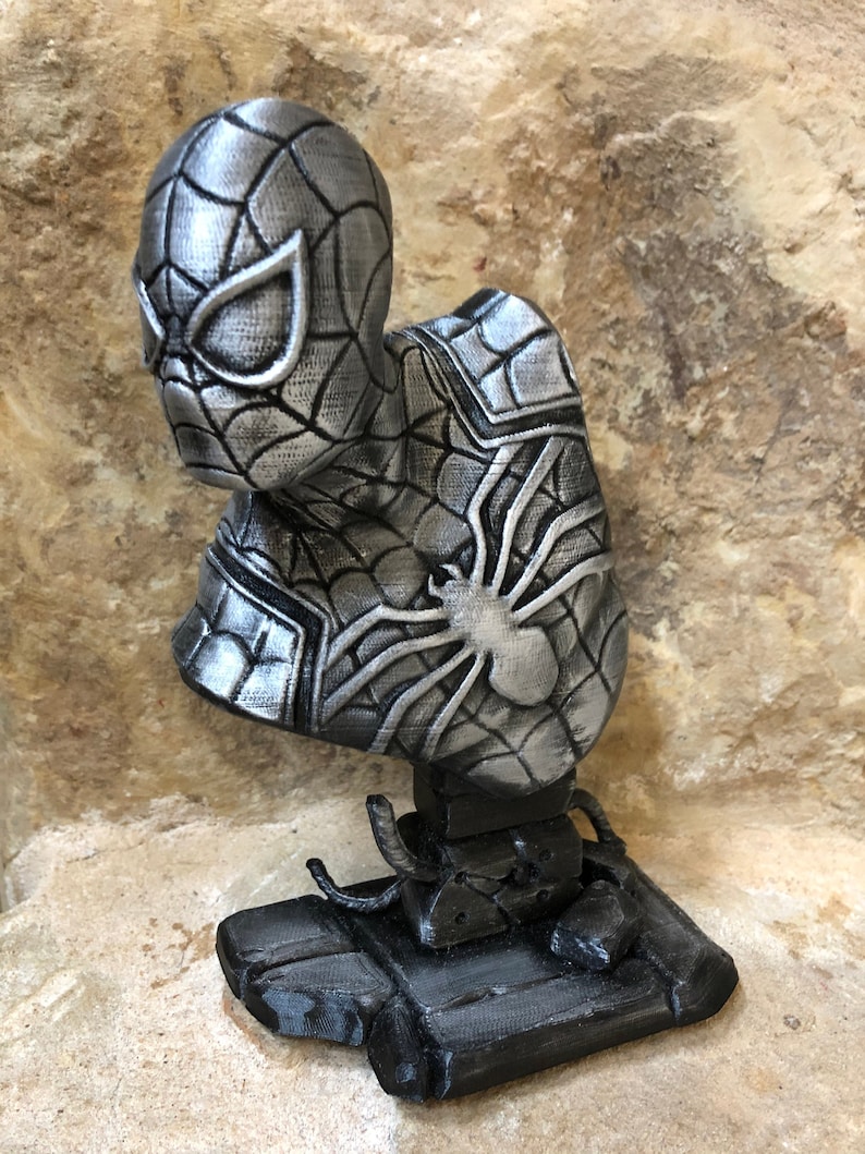 May include: A silver 3D printed bust of Spider-Man with a detailed web design. The bust is mounted on a black base.
