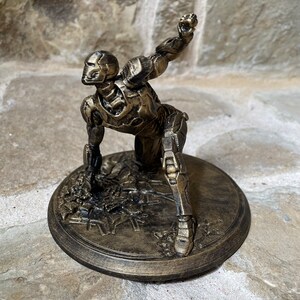 May include: A gold-painted figurine of a robot in a crouching pose, with one arm raised and a fist clenched. The figurine is standing on a round base with a textured surface.