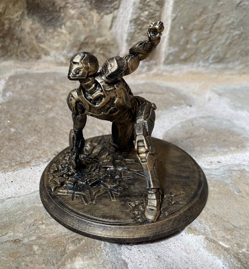 May include: A gold-painted figurine of a robot in a crouching pose, with one arm raised and a fist clenched. The figurine is standing on a round base with a textured surface.