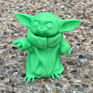 May include: A bright green figurine of a character with large ears and a robe. The figure is standing on a stone surface. The figurine is a popular character from a science fiction franchise.