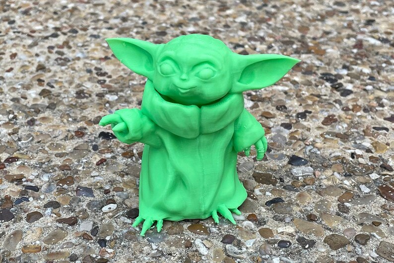 May include: A bright green figurine of a character with large ears and a robe. The figure is standing on a stone surface. The figurine is a popular character from a science fiction franchise.