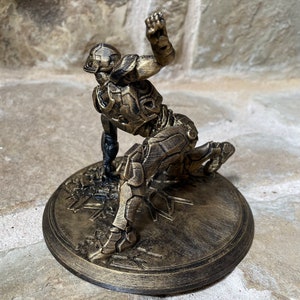 May include: A gold-coloured 3D printed figurine of a superhero in a crouching pose. The figurine is on a round base with a textured surface.