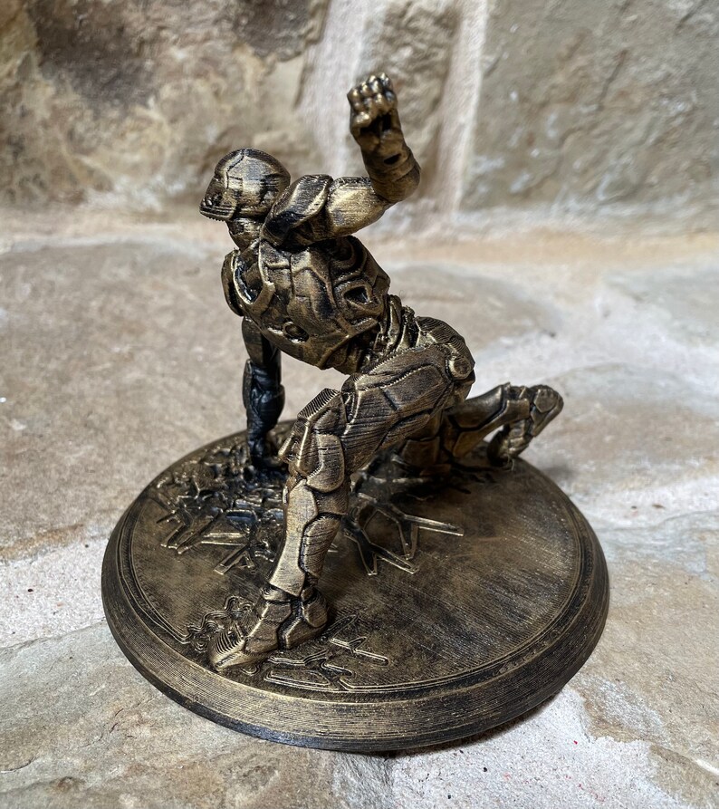 May include: A gold-coloured 3D printed figurine of a superhero in a crouching pose. The figurine is on a round base with a textured surface.