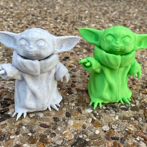 May include: Two figurines of a character with large ears and a robe. One is light gray with dark speckles, the other is bright green. Both have outstretched arms and are standing on a stone surface. The figures are approximately 4 inches tall.