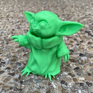 May include: A bright green figurine of a character with large ears and a hooded robe. The figure has outstretched arms and is standing on a textured surface. The figurine is a representation of a popular character from a science fiction franchise.