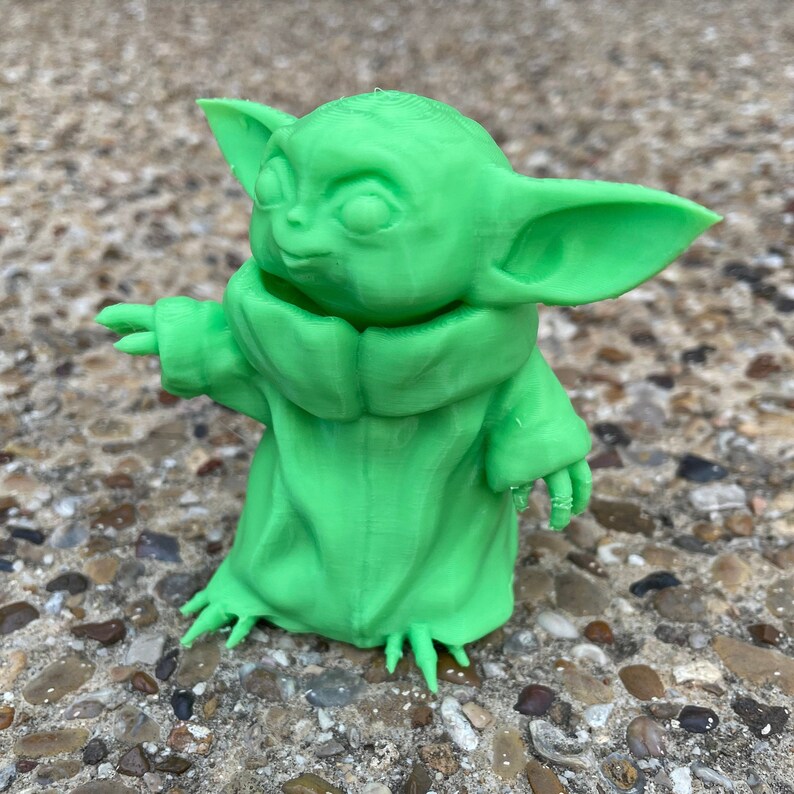 May include: A bright green figurine of a character with large ears and a hooded robe. The figure has outstretched arms and is standing on a textured surface. The figurine is a representation of a popular character from a science fiction franchise.