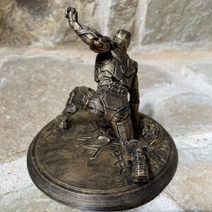 May include: A gold-coloured 3D printed figurine of a superhero in a kneeling pose. The figurine is on a round base with a textured surface.