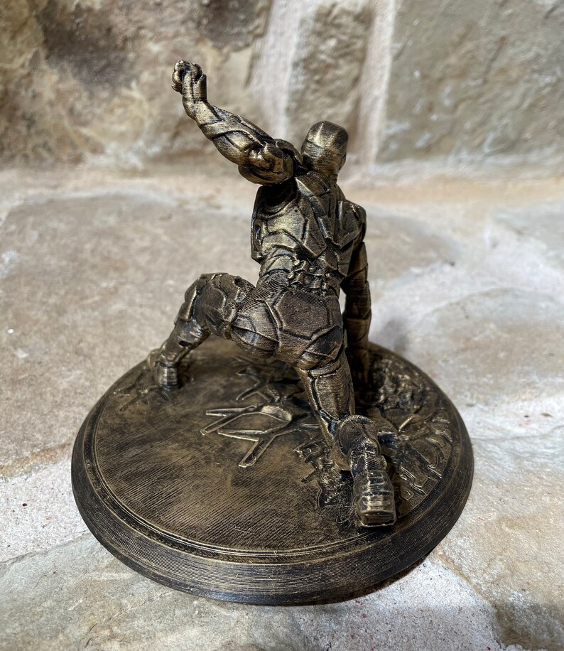 May include: A gold-coloured 3D printed figurine of a superhero in a kneeling pose. The figurine is on a round base with a textured surface.