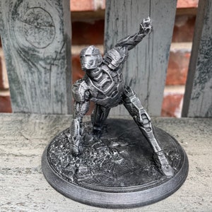 May include: A 3D printed figurine of a robot in a crouching pose. The robot is made of grey plastic and has a detailed design. The figurine is standing on a circular base.