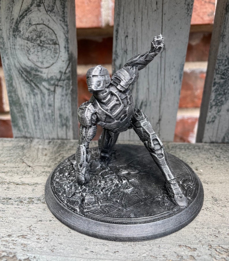 May include: A 3D printed figurine of a robot in a crouching pose. The robot is made of grey plastic and has a detailed design. The figurine is standing on a circular base.