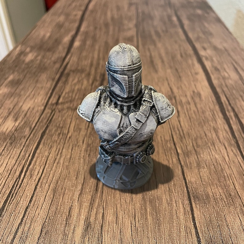 May include: A gray 3D printed bust of a Mandalorian character from the Star Wars franchise. The bust features a detailed helmet and armor, and is posed with a neutral expression.