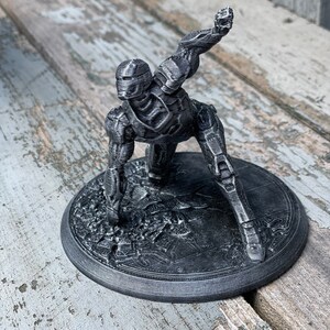 May include: A grey 3D printed figurine of a robot in a dynamic pose, standing on a circular base with a textured surface.