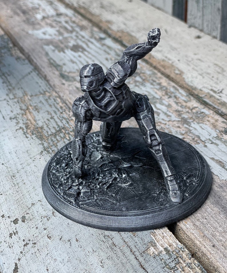 May include: A grey 3D printed figurine of a robot in a dynamic pose, standing on a circular base with a textured surface.