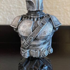 May include: A 3D printed bust of a Mandalorian character from the Star Wars universe. The bust is painted in a silver color and features a detailed helmet and armor. The bust is mounted on a black base.
