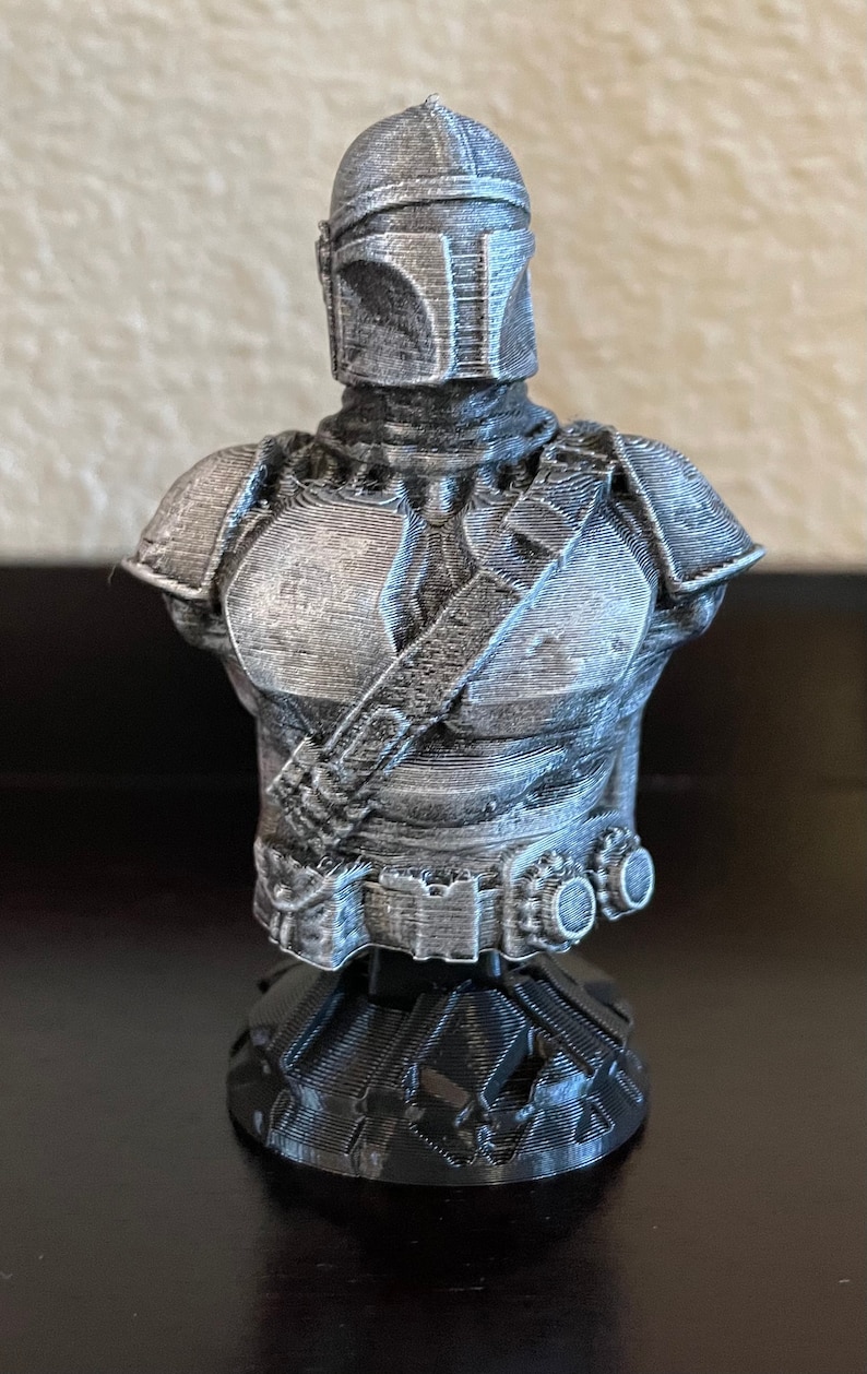 May include: A 3D printed bust of a Mandalorian character from the Star Wars universe. The bust is painted in a silver color and features a detailed helmet and armor. The bust is mounted on a black base.