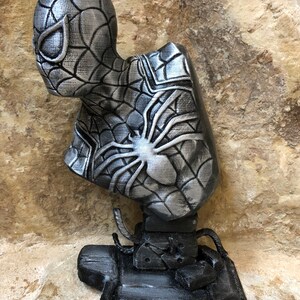 May include: A silver 3D printed bust of Spider-Man with a black web design. The bust is on a black base with web-like details.