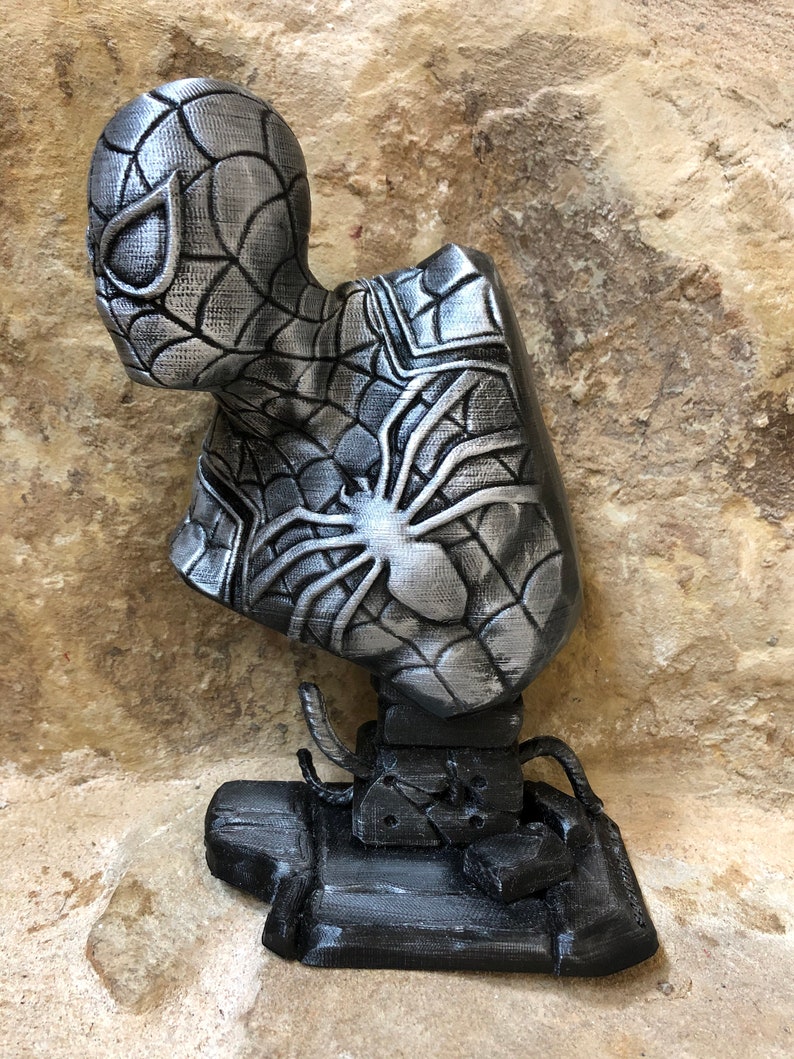 May include: A silver 3D printed bust of Spider-Man with a black web design. The bust is on a black base with web-like details.