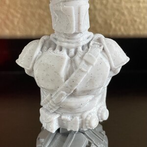 May include: A white 3D printed bust of a Mandalorian character from the Star Wars franchise. The bust features a detailed helmet and armor with a speckled texture. The character is holding a weapon in their right hand.