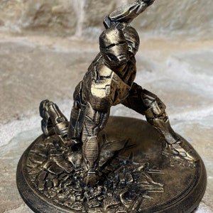 May include: A gold-coloured 3D printed figurine of a robot in a fighting pose. The robot is standing on a circular base with a textured surface.