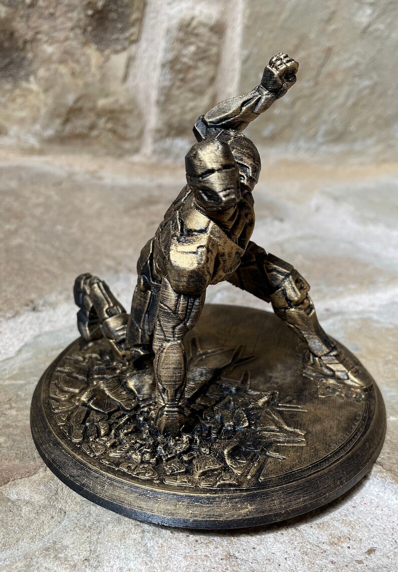 May include: A gold-coloured 3D printed figurine of a robot in a fighting pose. The robot is standing on a circular base with a textured surface.