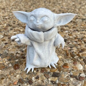 May include: A light gray figurine of a character with large ears, a small nose, and large eyes. The figurine is wearing a cloak and has small hands and feet. The figurine is speckled with small black dots.