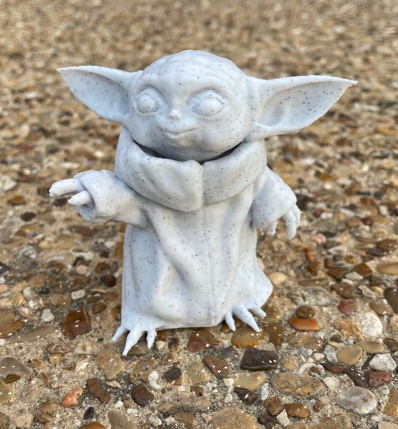 May include: A light gray figurine of a character with large ears, a small nose, and large eyes. The figurine is wearing a cloak and has small hands and feet. The figurine is speckled with small black dots.