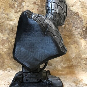 May include: A black and silver 3D printed bust of Spider-Man. The bust is posed with the head turned to the side and the body facing forward. The bust is mounted on a black base.