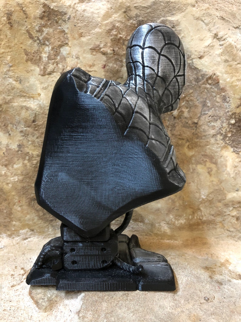 May include: A black and silver 3D printed bust of Spider-Man. The bust is posed with the head turned to the side and the body facing forward. The bust is mounted on a black base.