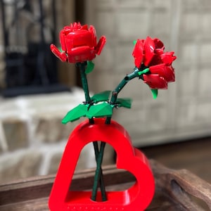 May include: A red heart-shaped vase holds two red rose flowers with green stems and leaves. The vase is on a wooden tray. The roses are made of plastic and appear to be a decorative item.