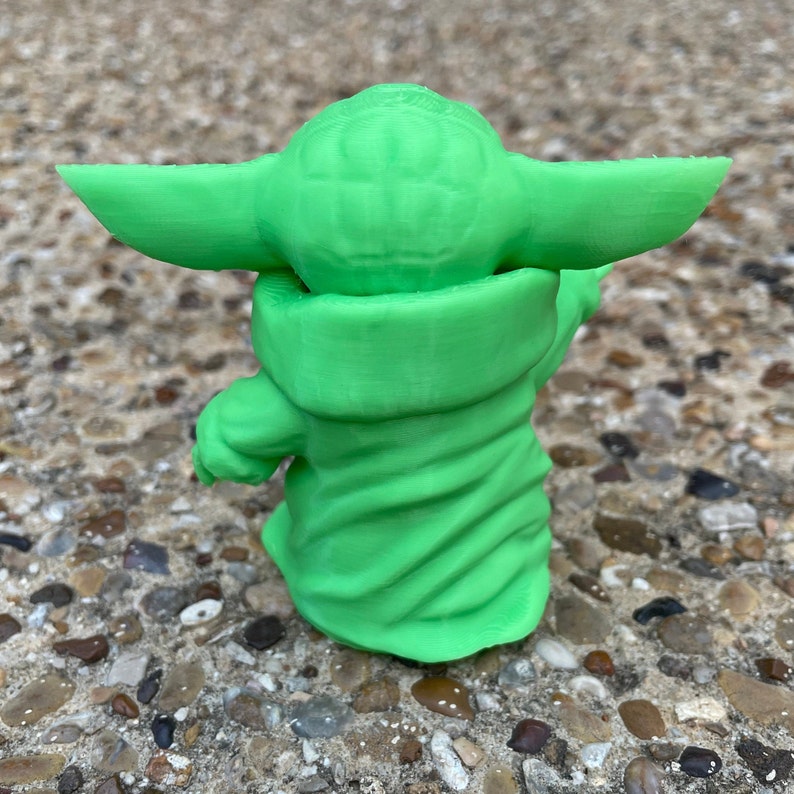 May include: A green 3D-printed figurine of a character with large ears and a robe. The figurine is standing on a textured surface. The figure is a light green color.