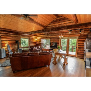 May include: Interior shot of a rustic cabin with exposed wooden beams and walls. A brown leather sofa sits in front of a window, and a wooden dining table is visible. The room has a cozy, natural aesthetic.