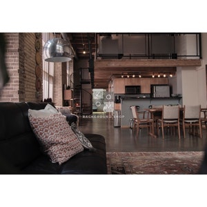 May include: An interior shot of a modern loft apartment with exposed brick, a spiral staircase, and a kitchen area. A black leather sofa with patterned pillows is in the foreground. The space is well-lit with natural light.