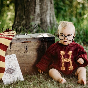 May include: A baby wearing a red and gold knitted sweater with a large 'H' on the front, sitting in a grassy area with a wooden chest and a lantern. The baby is wearing round glasses.