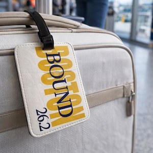 May include: A luggage tag with the words "BOSTON BOUND 26.2" in yellow and navy blue on a white background. The tag is attached to a light grey suitcase with a black strap. The image is taken in an airport setting.
