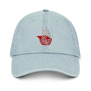 May include: A light blue denim baseball cap featuring an embroidered design of a white soft-serve ice cream cone with colorful sprinkles inside a red helmet-shaped cup. The red cup has the white text GO PHILS! embroidered on the front.