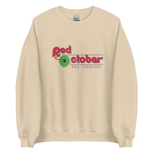 Puede incluir: Una sudadera beige con un estampado gráfico rojo y verde que dice "Red October" con las palabras "The Fightins" debajo.