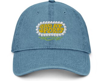May include: A light blue denim baseball cap with an embroidered design. The design features the words "BAD AT SPORTS" in yellow, outlined in white, and framed by a daisy-like border and green leaves.