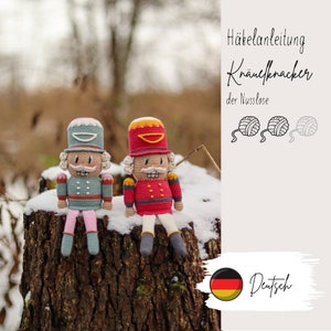 Könnte beinhalten: Zwei gehäkelte Nussknacker-Puppen, eine in Rot und eine in Blau, sitzen auf einem Baumstumpf. Die Puppen haben Wollknäuel und den Text "Häkelanleitung Knäuelknacker der Nusslose" und "Deutsch" im Hintergrund.