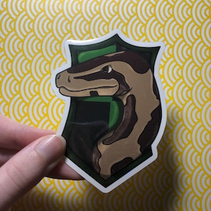 Può includere: Un adesivo raffigurante una testa di serpente all'interno di uno stemma verde e bianco. Il serpente è marrone e beige con segni marrone scuro. Lo stemma è posto su uno sfondo a motivi gialli e bianchi.