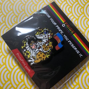 May include: A gecko enamel pin with a brown and yellow spotted pattern, holding a flag with the colors of the non-binary pride flag. The pin is on a black backing card with the text "Show your pride. You deserve it." and "Pride Pins."