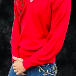 Könnte beinhalten: Ein leuchtend roter V-Ausschnitt-Pullover mit einem Erdbeer-Aufnäher auf der Brust, getragen über einem hellrosa Hemd mit Kragen und einer blauen Jeans. Ein mehrlagiger goldfarbener Kettengürtel mit kleinen Anhängern hängt an der Jeans.