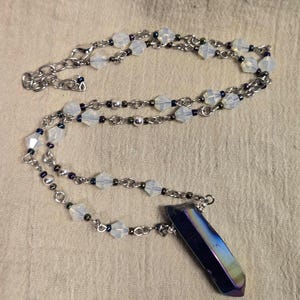 May include: A necklace featuring a dark iridescent purple crystal pendant suspended from a silver-toned chain. The chain is accented with translucent white faceted beads and small dark metallic spacer beads, finished with a lobster claw clasp.