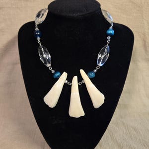 May include: A necklace featuring three off-white, tooth-shaped pendants suspended from a silver-toned chain. The chain is accented with alternating clear, faceted, oval-shaped beads and round, dark blue, metallic-finish beads.