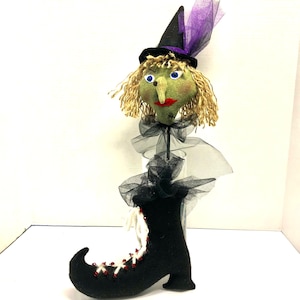 May include: A green felt witch doll with a black hat and purple tulle, standing in a glass vase with a black felt boot.